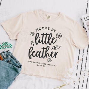 an ivory comfort colors 3023cl t-shirt with bold white text that says 'Mocks by Little Feather – Real People. Real Clothes. Zero AI.' surrounded by doodle-style flowers and feathers. Trendy Etsy-style mockup featuring real model photography for print-on-demand sellers.