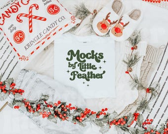 Könnte beinhalten: Ein weißes T-Shirt mit dunkelgrünem Text Mocks by Little Feather, umgeben von einer Süßigkeitentüte mit der Aufschrift Kringle Candy Co., Rentier-Hausschuhen, weißen Strickstrümpfen und einer Girlande aus roten Beeren und Tannennadeln.