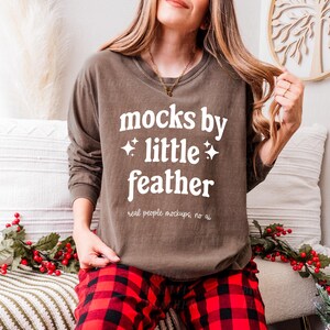 May include: A brown long-sleeved shirt with the text "mocks by little feather" and the words "real people mockups, no ai" printed on the front. The shirt is being worn by a person with long brown hair and red and black checked trousers.