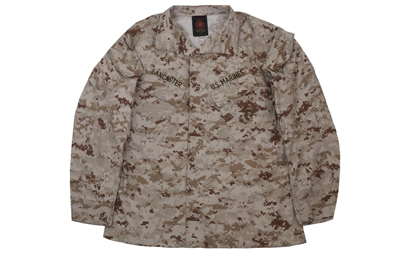 Puede incluir: Una camisa de uniforme militar de camuflaje marr&oacute;n y beige con el texto "U.S. Marines" y "LANCASTER" en la parte delantera.