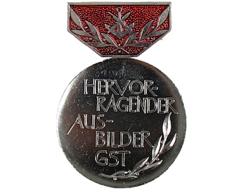 May include: A silver medal with a red ribbon. The medal features a laurel wreath and the text "HERVOR-RAGENDER AUS-BILDER GST".