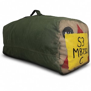 May include: A rectangular olive green canvas duffel bag with a top handle. The end panel features a large yellow rectangular patch with the black hand-painted text S3 M8731 C, alongside partial red and black circular markings on the fabric.