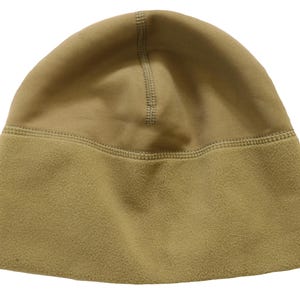 May include: A tan beanie cap constructed from two distinct fabric textures. The upper portion features a smooth, solid finish, while the lower band is crafted from a soft, brushed fleece material. A horizontal seam connects the two sections.