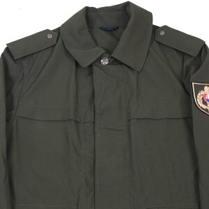 May include: A dark green military jacket with a patch on the right sleeve featuring a gold emblem with a shield and a crown. The jacket has a button-up front and a collar.
