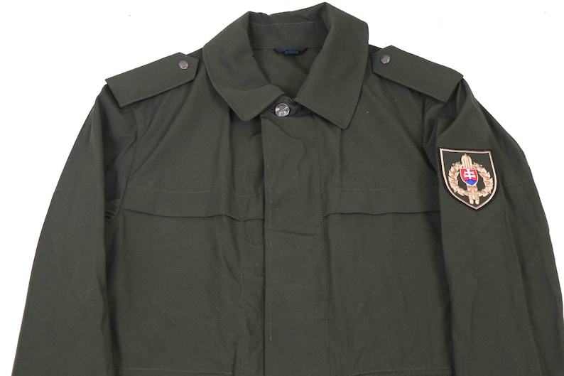 May include: A dark green military jacket with a patch on the right sleeve featuring a gold emblem with a shield and a crown. The jacket has a button-up front and a collar.