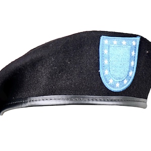 May include: A black wool beret featuring a black leather trim along the bottom edge. Attached to the front is a light blue embroidered patch shaped like a shield, bordered by a series of small white stars arranged in a circular pattern.