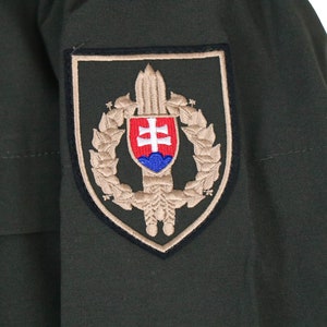 May include: A green military jacket with a tan patch on the sleeve. The patch features a shield with a sword, a wreath, and the Slovakian coat of arms.
