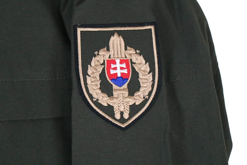 May include: A green military jacket with a tan patch on the sleeve. The patch features a shield with a sword, a wreath, and the Slovakian coat of arms.