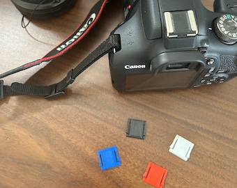 May include: A black Canon camera with a lens attached and a black strap. The camera is sitting on a wooden surface with four small colored clips: black, blue, white, and red.