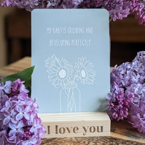 May include: A light blue rectangular card featuring a white line drawing of two sunflowers in a jar and the text "MY BABY IS GROWING AND DEVELOPING PERFECTLY" resting on a light wood block stand with the engraved text "I love you".