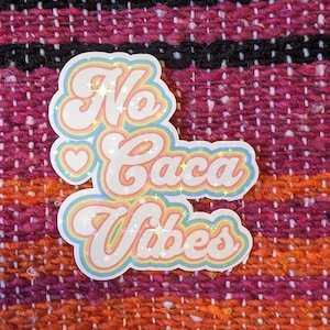 May include: A sticker with the words "No Caca Vibes" in a pastel rainbow design with a heart. The sticker has a holographic, glittery effect and is set against a textured, colorful background.