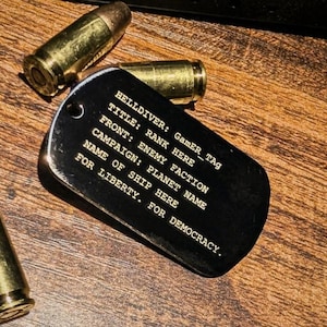 May include: A black metal dog tag with gold-colored engraved text reading HELLDIVER: GamER_Tag, TITLE: RANK HERE, FRONT: ENEMY FACTION, CAMPAIGN: PLANET NAME, NAME OF SHIP HERE, FOR LIBERTY. FOR DEMOCRACY. Three brass-colored bullet casings surround the tag.