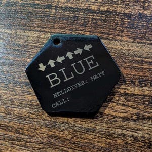 May include: A black, hexagonal tag with the word "BLUE" in large, silver letters. Above the word are four arrows pointing up and to the left. Below the word are the words "HELLDIVER: MATT" and "CALL:" in smaller silver text.