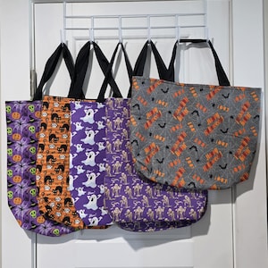 May include: Five fabric tote bags with black handles hanging on a white rack. The bags feature Halloween patterns including purple with pumpkins, orange with black cats, purple with white ghosts, purple with mummies, and gray with bats and text.