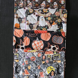 May include: A stack of four rectangular quilted fabric pot holders featuring various Halloween-themed prints. The patterns include gnomes with pumpkins, ghosts with rainbows and flowers, orange pumpkins on a black background, and a spooky haunted house scene.