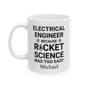以下が含まれることがあります： 「ELECTRICAL ENGINEER BECAUSE ROCKET SCIENCE WAS TOO EASY」の黒い文字がプリントされた白いセラミックマグ。ロケットのグラフィックが「ROCKET」の下に配置されています。「Michael」という名前が下に印刷されています。コーヒーマグ。