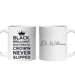 May include: Two white ceramic mugs. One mug features the text "BLACK. BRILLIANT. DOCTORATE. CROWN NEVER SLIPPED" with a crown graphic. The other mug has the name "Dr. Williams" in cursive.