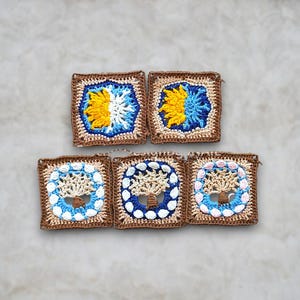 Op de afbeelding: Vijf handgemaakte gehaakte vierkanten. De vierkanten hebben een bruine rand met een centraal ontwerp. Twee vierkanten hebben een geel en wit bloemmotief, terwijl de andere drie een boomontwerp hebben met blauwe en witte accenten. De vierkanten zijn gerangschikt op een lichtgrijze ondergrond.