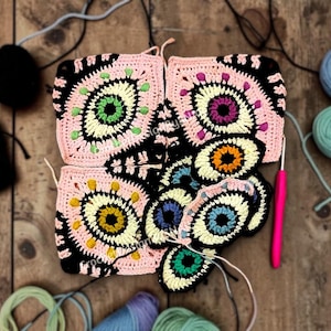 May include: A collection of colorful crochet eye-shaped squares and circles in various stages of completion. The squares feature pink, black, and cream colors with green, yellow, and purple accents. A pink crochet hook and yarn are also visible.