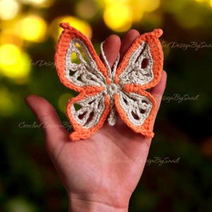 May include: A handmade crochet butterfly in orange and cream colors. The butterfly is held in a hand, showcasing its intricate openwork design. The wings have an orange border and cream-colored interior. The background is blurred with green and yellow hues.
