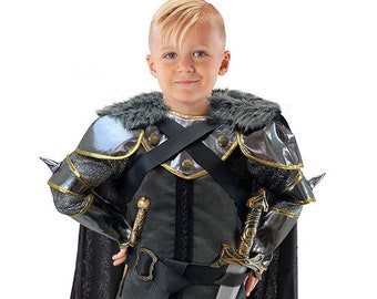 May include: A child wearing a black and silver knight costume with a faux fur collar, holding a silver sword and wearing a black cape. A silver helmet with a black interior is on the floor.
