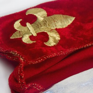 May include: A red velvet fabric with a gold embroidered fleur-de-lis design. The fabric has a gold stitched edge.
