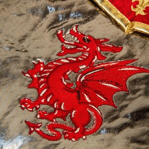 May include: A red embroidered dragon with white accents on a silver background. The dragon is facing right and has its wings spread out.