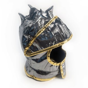 May include: A silver and gold knight helmet costume for kids. The helmet has a pointed top and a face mask with a small opening for the eyes.