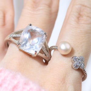 Puede incluir: Dos anillos de plata con piedras claras y blancas. El anillo en el dedo índice tiene una piedra grande, clara y de forma cuadrada. El anillo en el dedo medio tiene una pequeña perla blanca y un diseño en forma de trébol con piedras blancas más pequeñas.