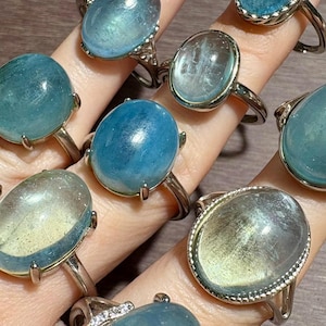 Op de afbeelding: Een collectie aquamarijn edelsteenringen in diverse vormen en zettingen. De ringen hebben ovale en ronde stenen in blauwe en groene tinten, gezet in zilveren banden. Sommige ringen hebben decoratieve details.