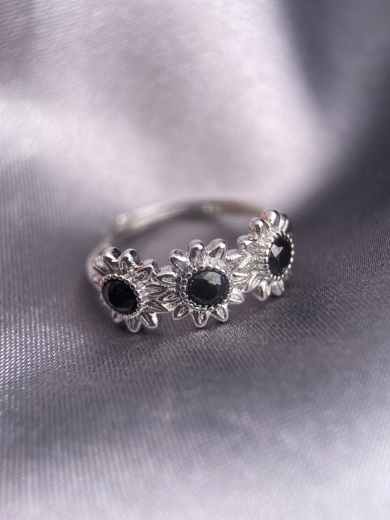 May include: A silver ring with three black gemstones set in a sunflower design.