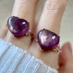 May include: Two rings featuring heart-shaped, purple gemstones. The rings have a rose gold band with small decorative details around the gemstone. The gemstones have a shimmering, reflective appearance. The rings are displayed on a person's fingers.