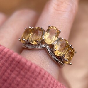 May include: A silver ring with a row of five oval-shaped yellow gemstones. The ring has a V-shaped design in the center.