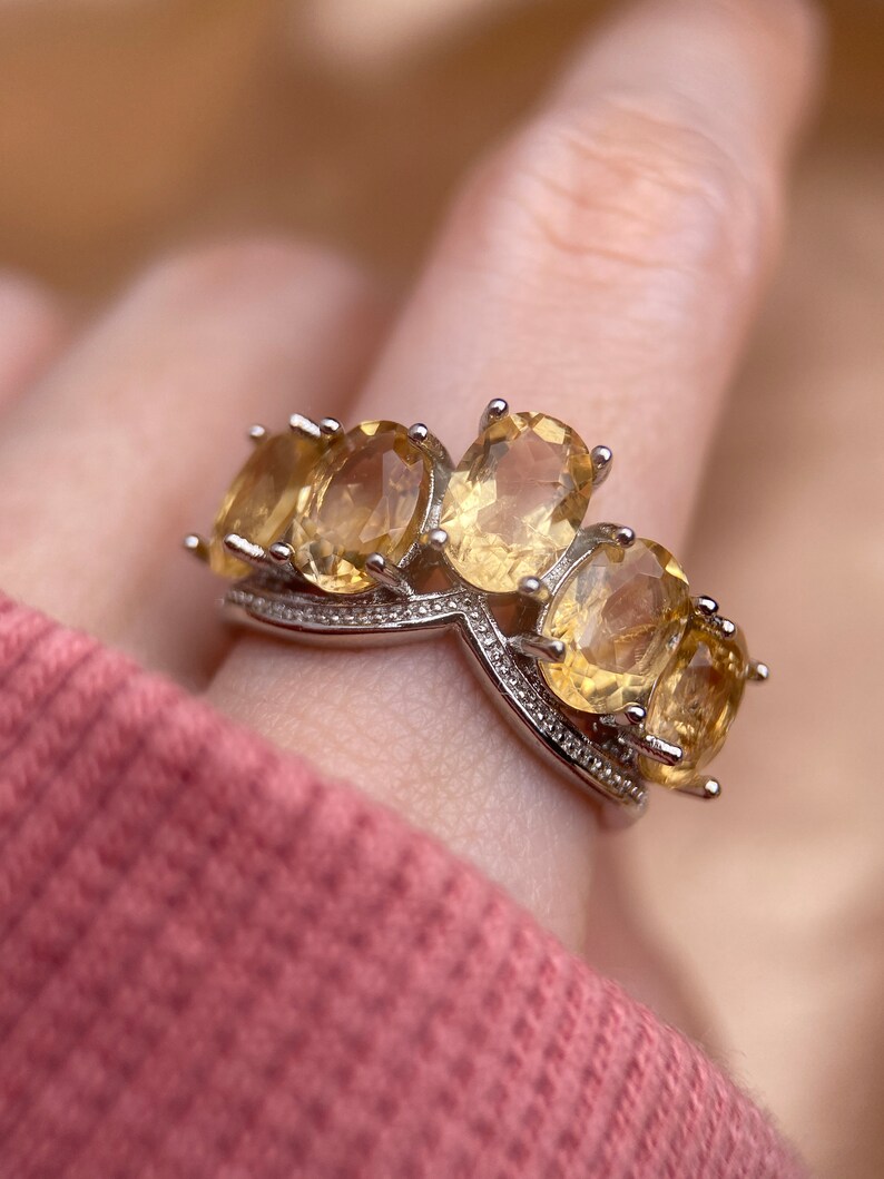May include: A silver ring with a row of five oval-shaped yellow gemstones. The ring has a V-shaped design in the center.