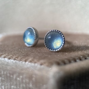 May include: Two stud earrings featuring round, iridescent blue gemstones set in silver-coloured metal. Each earring has a beaded edge. The earrings are displayed on a soft, tan surface. The gemstones have a subtle, glowing effect.