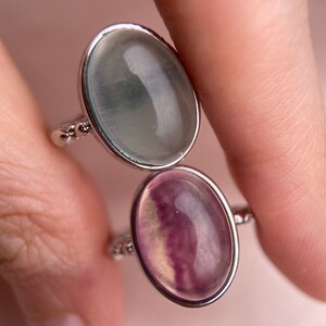 May include: Two oval-shaped rings with silver settings. The top ring features a light blue gemstone, while the bottom ring has a purple and yellow gemstone.