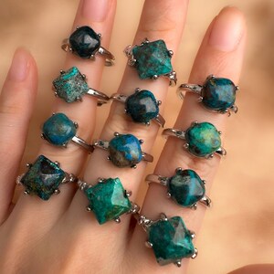 May include: A hand model wearing a set of 12 rings with different shades of green and blue gemstone settings. The rings are all silver and have a square or star-shaped setting.