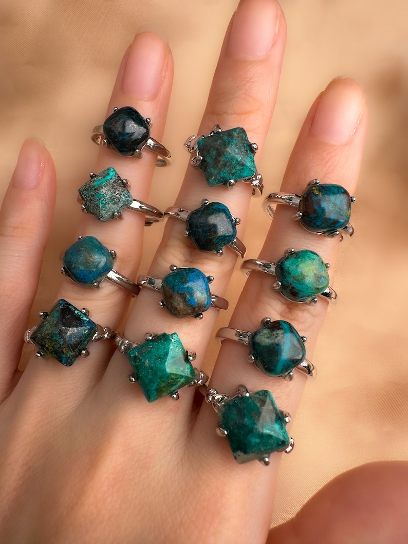 May include: A hand model wearing a set of 12 rings with different shades of green and blue gemstone settings. The rings are all silver and have a square or star-shaped setting.