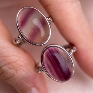 May include: Two silver rings with oval-shaped purple and white fluorite gemstones. The rings are set with small silver beads around the band.