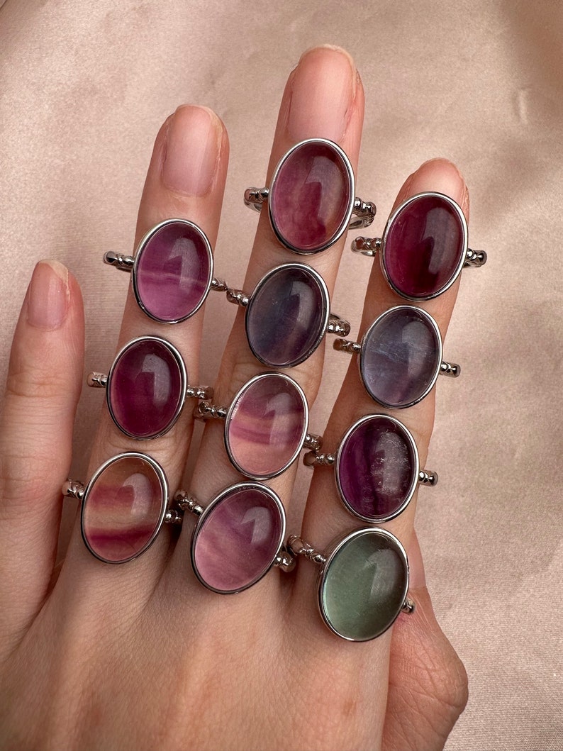 May include: A collection of oval-shaped rings with different colored fluorite stones. The rings are silver and have a beaded band.