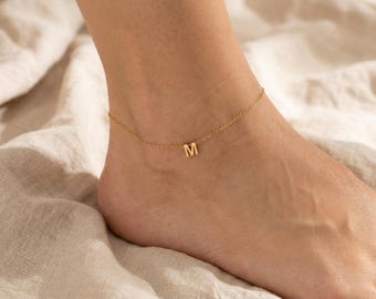 Puede incluir: Una tobillera de tono dorado que presenta una cadena delicada y un dije con la letra mayúscula M. La pieza está diseñada con una estructura de eslabones simples, mostrando un acabado metálico pulido que se ajusta alrededor del tobillo.