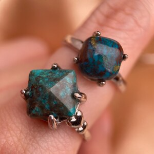 May include: Two silver rings with turquoise gemstones. The rings are stacked on a finger. The gemstones are a dark blue-green color with brown and gold flecks.