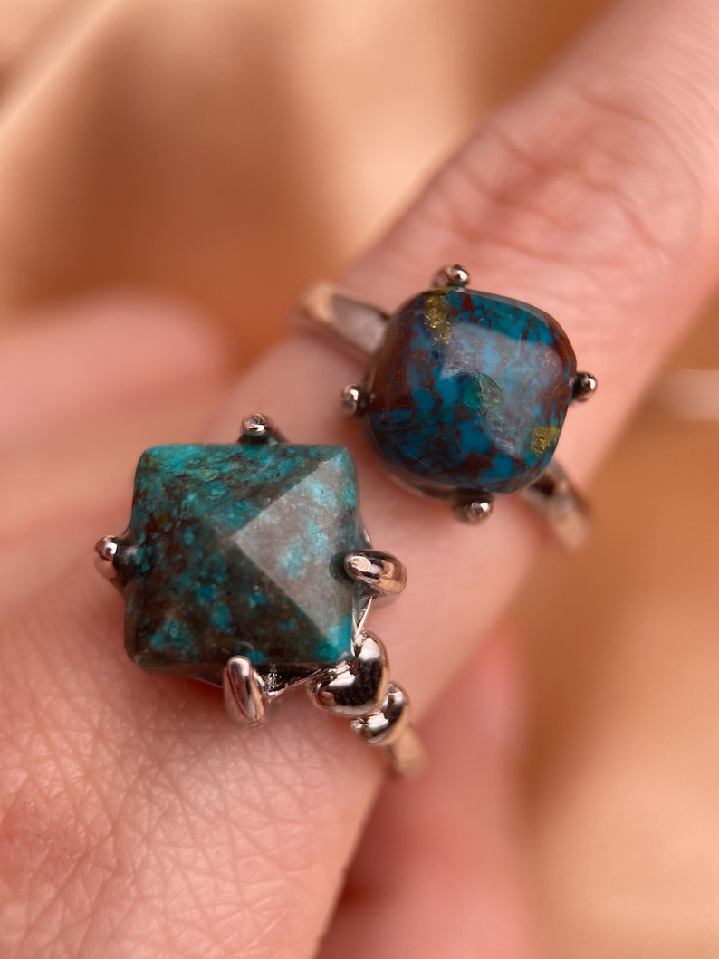 May include: Two silver rings with turquoise gemstones. The rings are stacked on a finger. The gemstones are a dark blue-green color with brown and gold flecks.