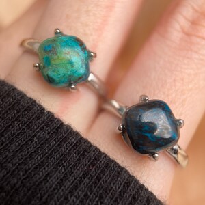 May include: Two silver rings with square-shaped turquoise gemstones. The top ring has a green and blue gemstone, while the bottom ring has a dark blue gemstone.