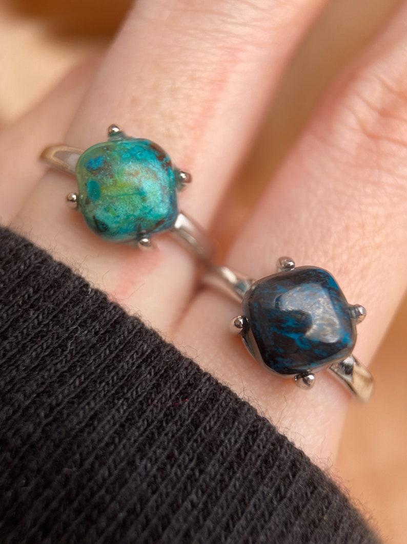 May include: Two silver rings with square-shaped turquoise gemstones. The top ring has a green and blue gemstone, while the bottom ring has a dark blue gemstone.