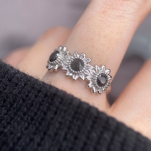 May include: Silver ring with three black sunflower designs. Each sunflower has a black gemstone in the center.