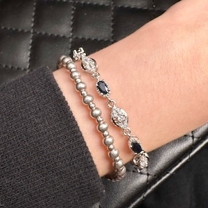 May include: Two silver bracelets with black gemstones and clear crystals. The bracelets are stacked on a person's wrist.