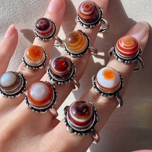 May include: A collection of gemstone rings with silver-colored bands. Each ring features a round, polished stone in shades of brown, orange, and white, set within a decorative bezel. The rings are displayed on fingers.