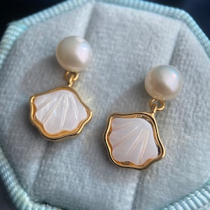 Puede incluir: Pendientes dorados con un diseño de concha blanca y una perla. Los pendientes tienen un borde dorado y están sobre un fondo de terciopelo azul claro.