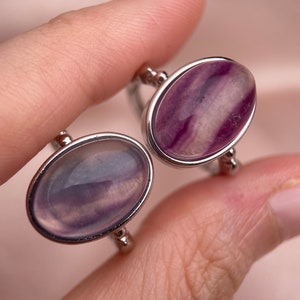 May include: Two silver rings with oval-shaped purple and white fluorite stones. The rings are on a finger.